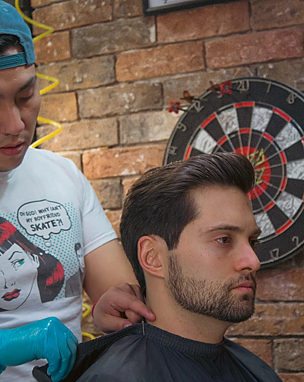 Barbero con guantes acomoda la capa de un cliente mientras revisa el perfil del corte, frente a una diana de dardos