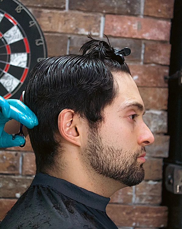 Cliente de perfil con cabello largo peinado hacia atrás y barba recortada; el barbero con guante azul termina el corte