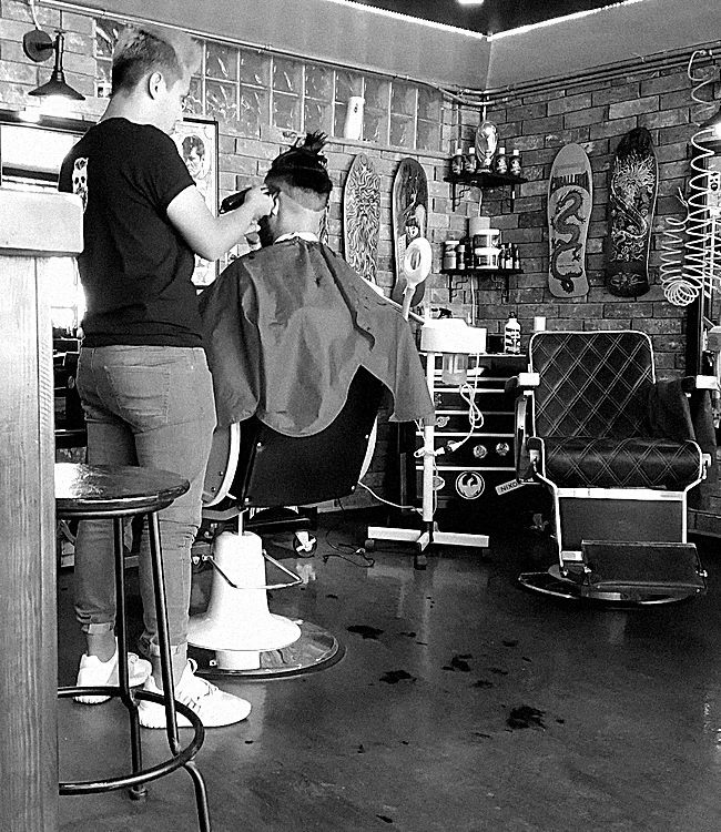 Interior de la barbería: un barbero corta el cabello junto a sillones de capitoné negro, patinetas colgadas en el muro de ladrillo y una manguera de aire en espiral