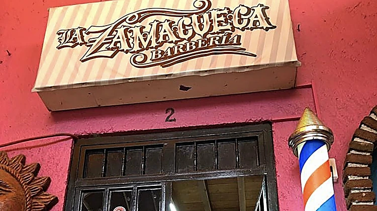 Letrero de madera clara con la leyenda La Zamacueca Barbería sobre una pared rosa, con un poste de barbero rojo, blanco y azul junto a la puerta